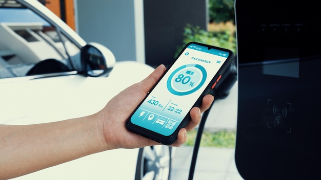A person using a mobile app to control and monitor their EV charging station. The app interface shows charging status, energy usage, and scheduling options. The charging station is visible in the background, connected to an electric car.