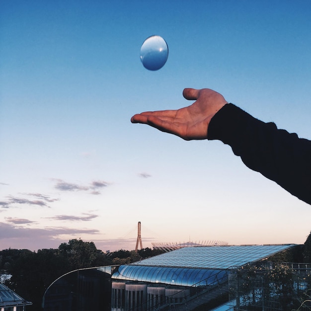 A close-up of a hand holding a small solar panel, with a cityscape in the blurred background. The image symbolizes the potential of individual contributions to the broader adoption of renewable energy in urban environments.
